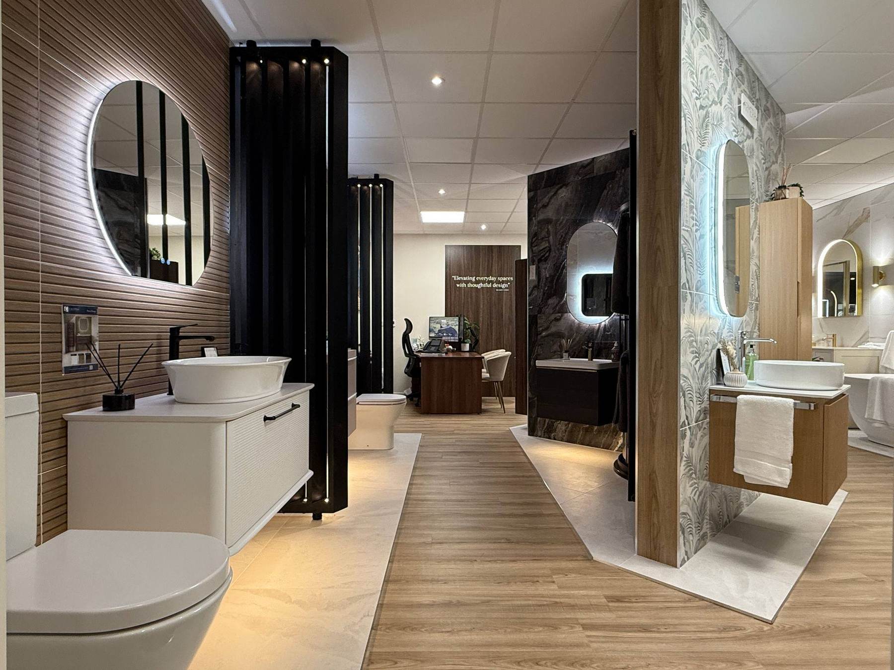 A Bath Works showroom aisle with vignettes side by side: a fluted timber wall with a backlit round mirror over a countertop basin, tall black radiators, a dark marble-effect bay with a lit oval mirror, and a botanical-print wall with an oak vanity and a white towel.