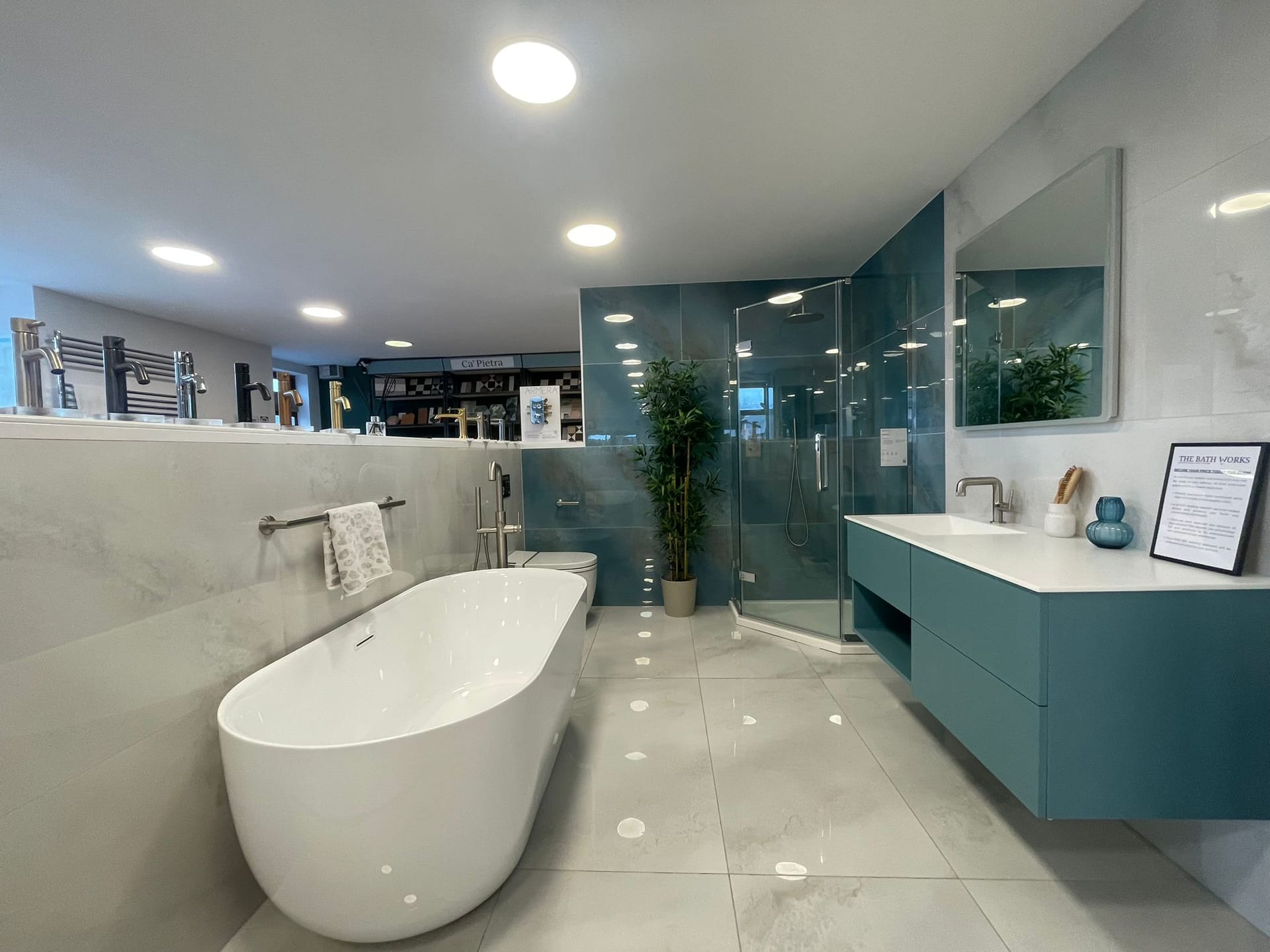 A room-set display in the New Milton showroom: a white oval freestanding bath on grey marble-effect tile, a wall-hung teal vanity with a white basin top and a framed card on it, a mirror above, a corner glass shower enclosure against teal tiling, a tall plant, and further displays lit by ceiling spots beyond.