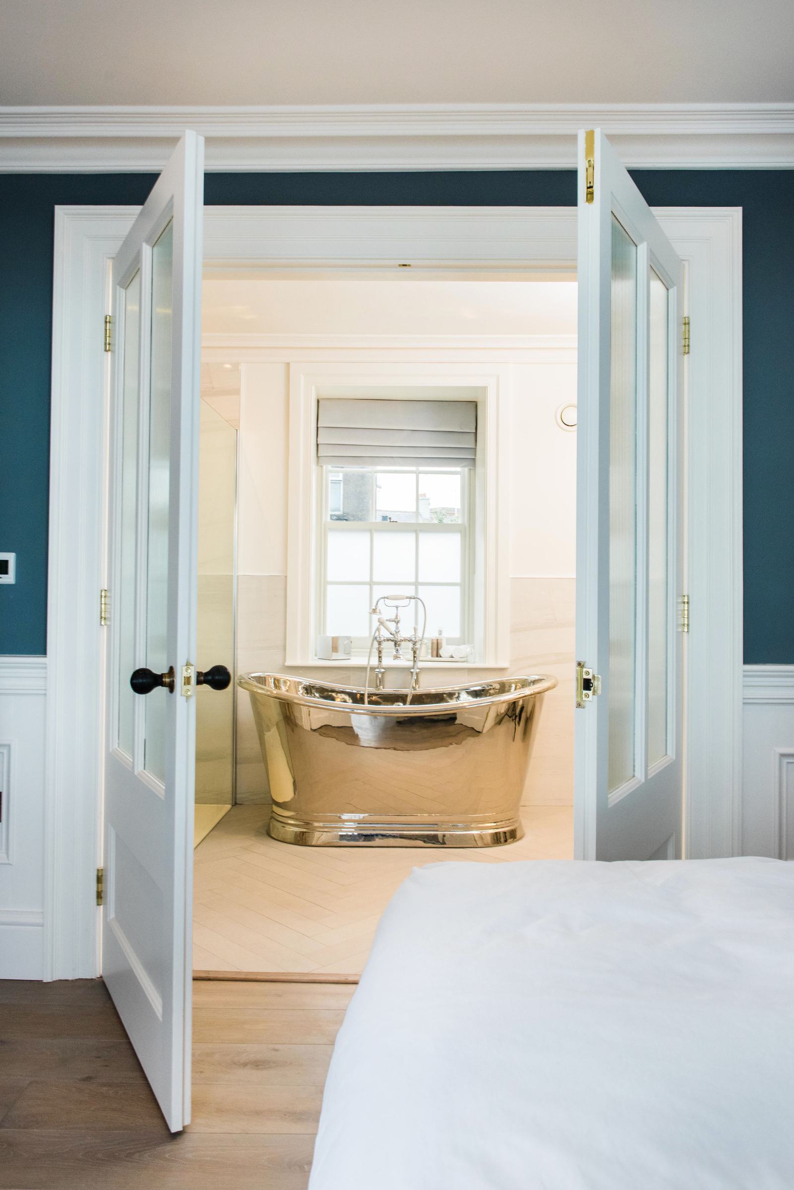 A view through open panelled double doors from a bedroom into an en-suite, a polished brass roll-top bath beneath a sash window.