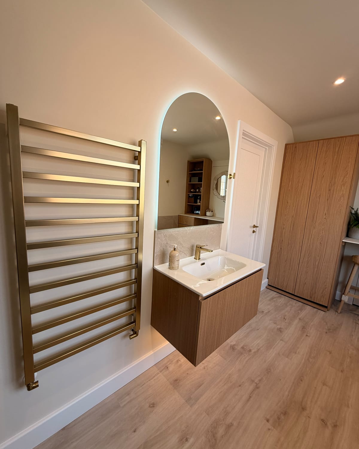 A brushed brass ladder towel rail beside a wall-hung fluted oak vanity with a white basin and a brushed brass mixer, under a backlit arched mirror, with a fluted white door and an oak wardrobe beyond.