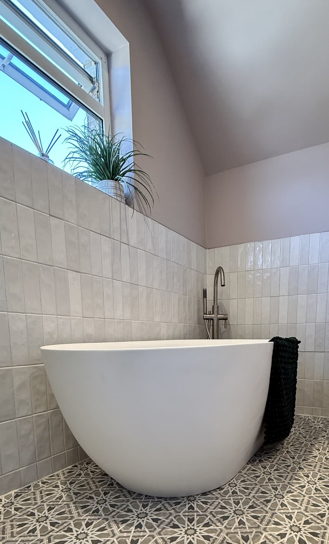 A matt white freestanding bath in the corner under a sloping ceiling and a high window, filled by a brushed nickel floor-standing tap with a handshower, against pale stacked tile with a patterned star-motif floor.