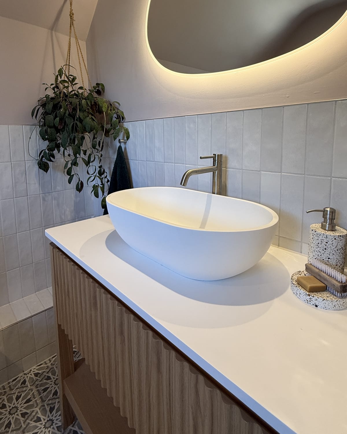A freestanding fluted oak washstand with an oval matt white countertop basin and a brushed nickel mixer, lit by the warm halo of a pebble-shaped mirror, with a trailing plant beside it and star-pattern floor tile below.