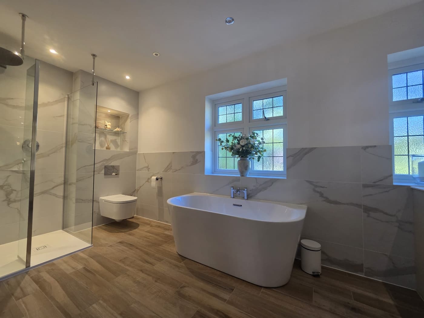 A white freestanding bath with a chrome deck-mounted filler below a window of small obscured-glass panes, beside a walk-in shower with a ceiling-mounted rain head, marble-effect tile, a lit niche, a wall-hung WC and a wood-effect plank floor.
