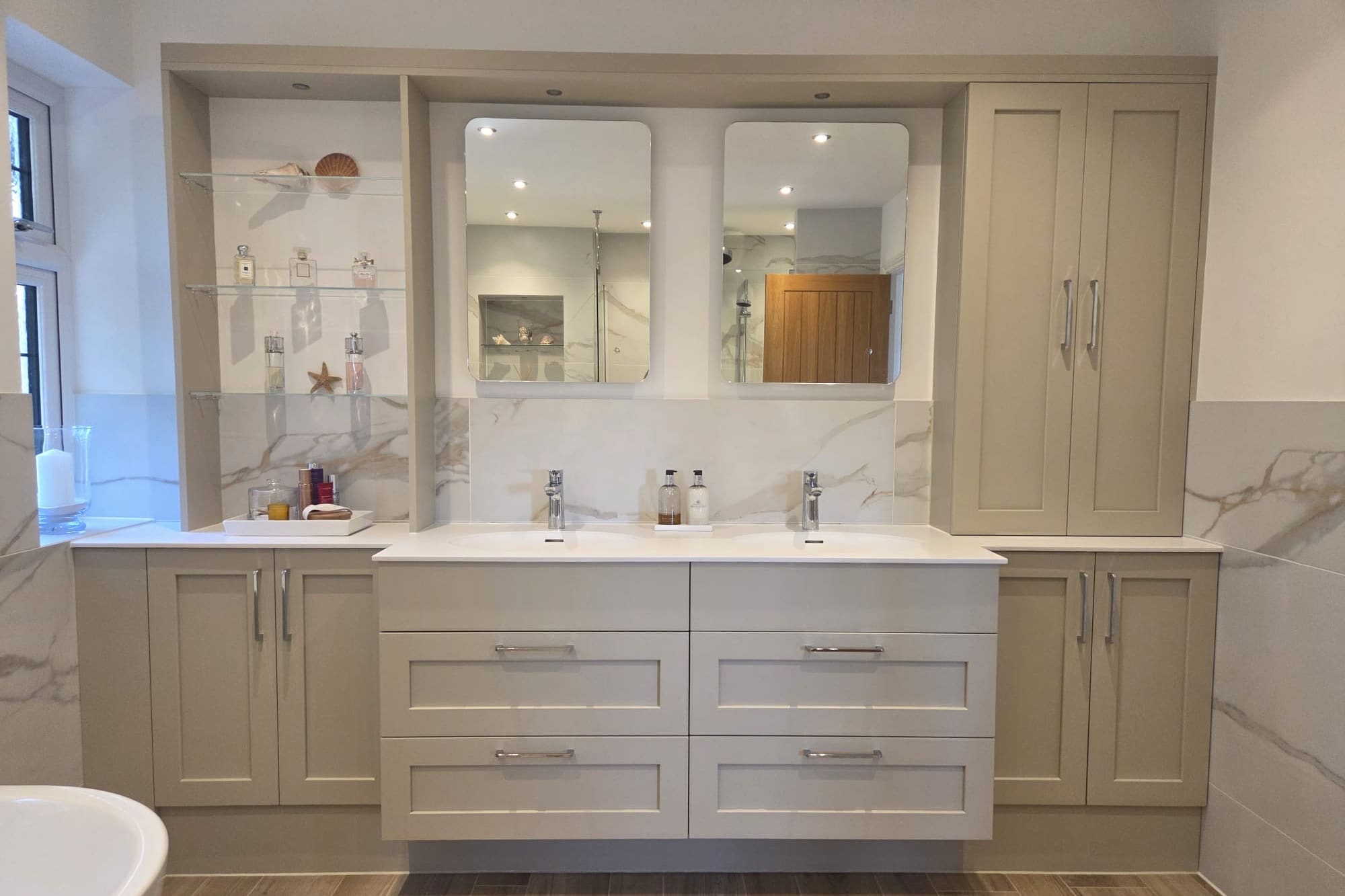 A full wall of taupe fitted bathroom furniture: a wall-hung double vanity with two moulded basins and chrome mixers under two frameless mirrors, flanked by cupboards and a glass-shelved display alcove, against large-format marble-effect porcelain.