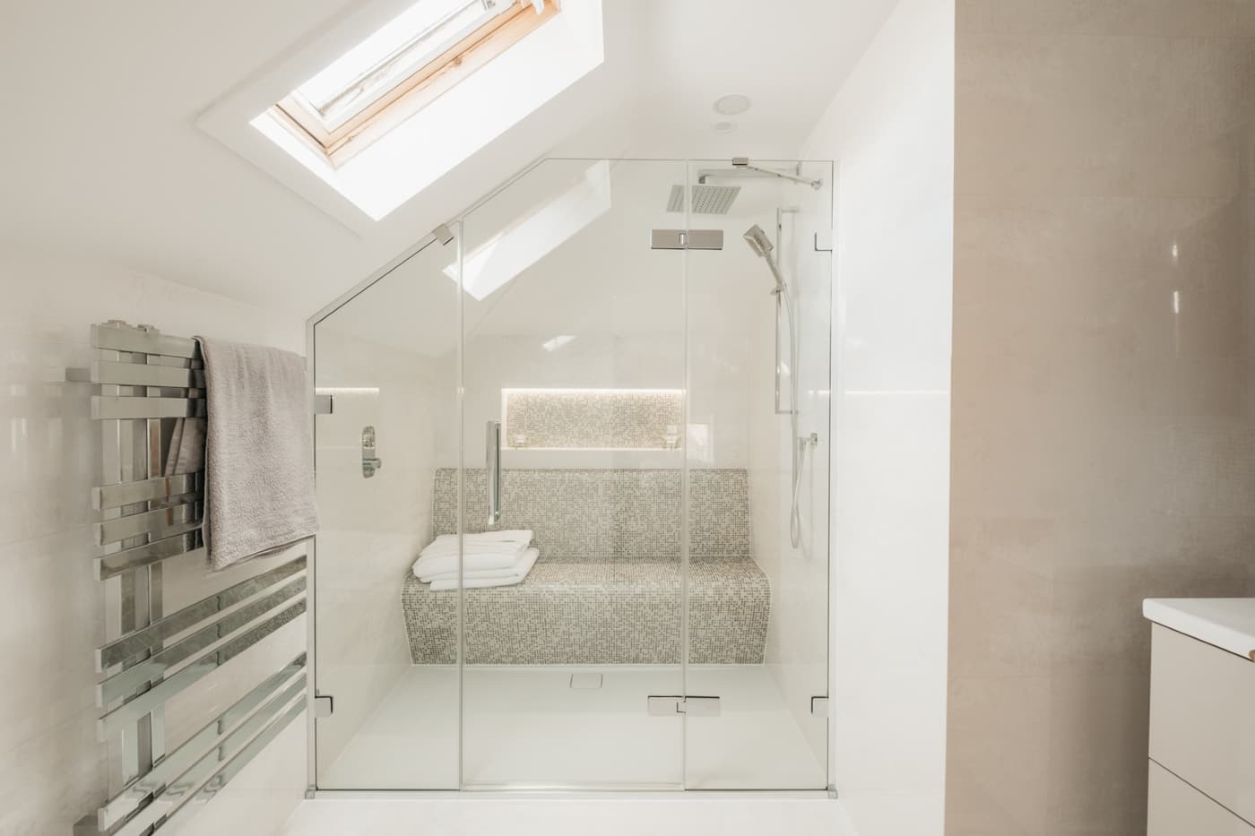 A glazed steam and shower enclosure built into a loft slope under a roof window, with a full-width mosaic bench, a lit mosaic niche and a chrome towel radiator alongside.