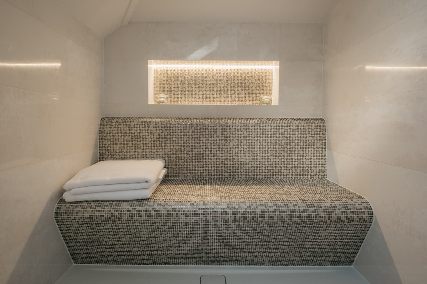 A contoured mosaic-tiled bench with a curved back and folded white towels, below a long backlit mosaic niche.