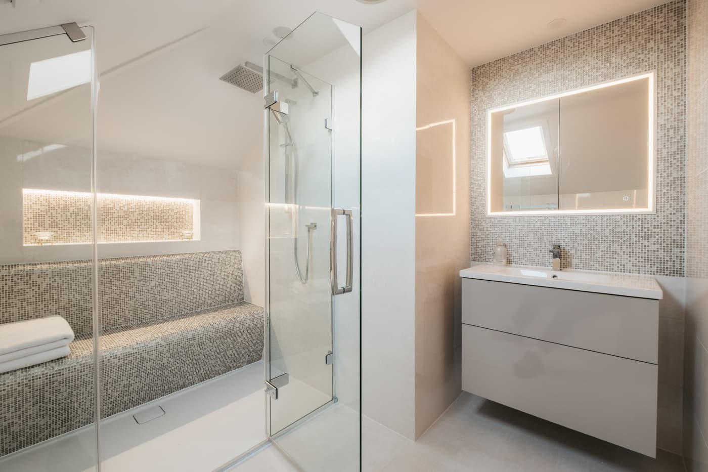 A white gloss wall-hung vanity with a wide basin and a backlit mirrored cabinet, set against a mosaic tiled wall, with the glazed shower enclosure to the left.