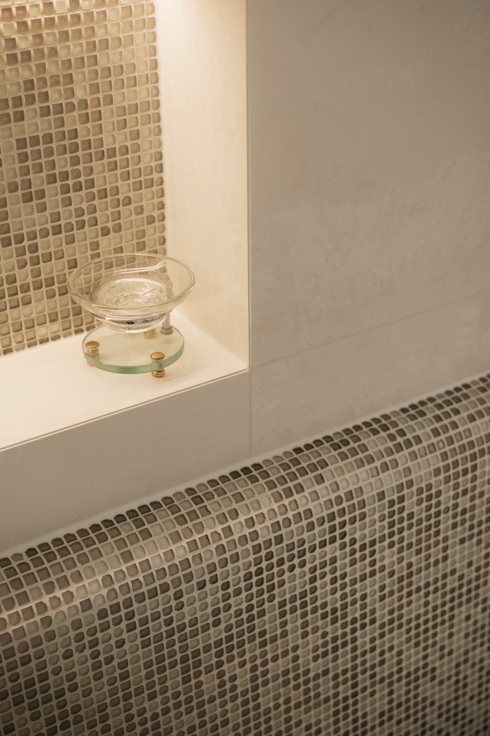 A lit mosaic-lined niche holding a clear glass soap dish on a small round glass disc raised on brass feet, above the curved mosaic edge of the shower bench.