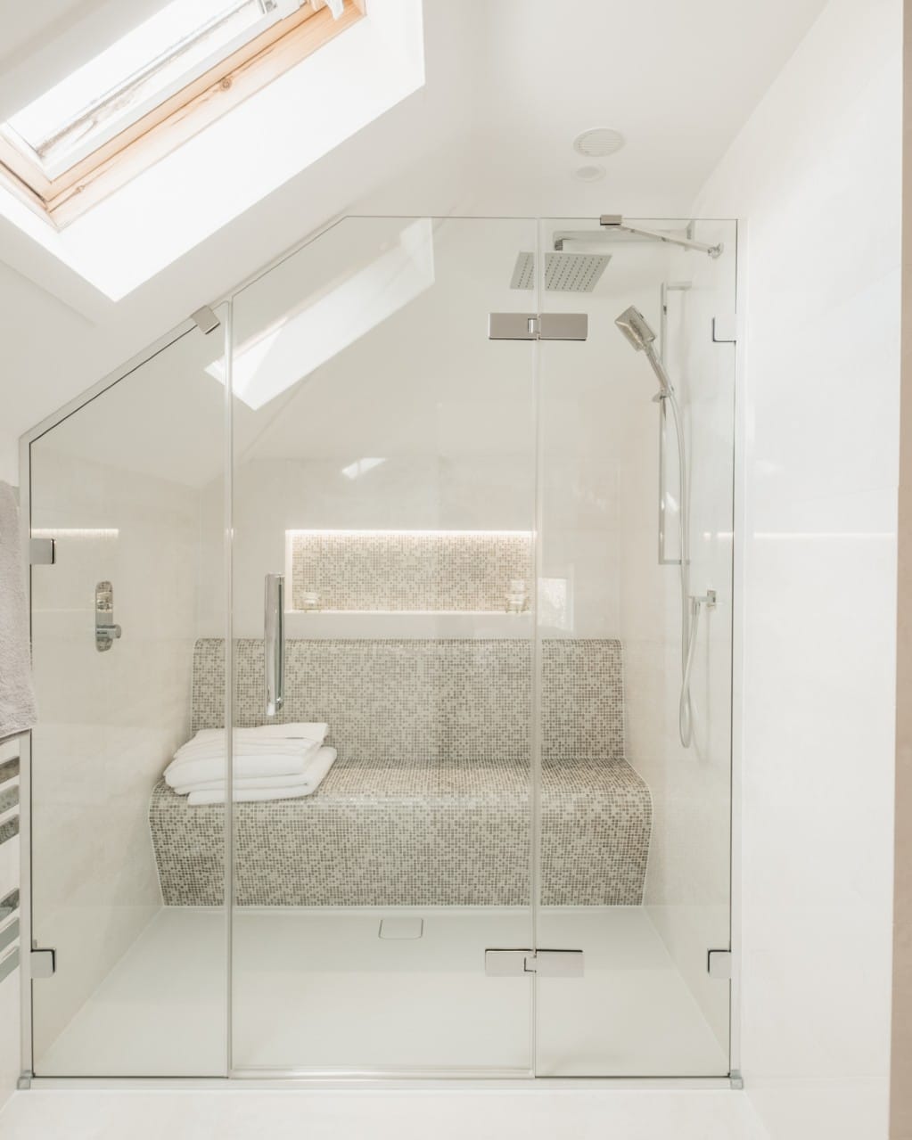 A frameless glass shower enclosure in a loft, holding a contoured mosaic-tiled bench with folded white towels and a backlit mosaic niche, with a square chrome rain head and handshower, lit by a rooflight in the pitched ceiling.