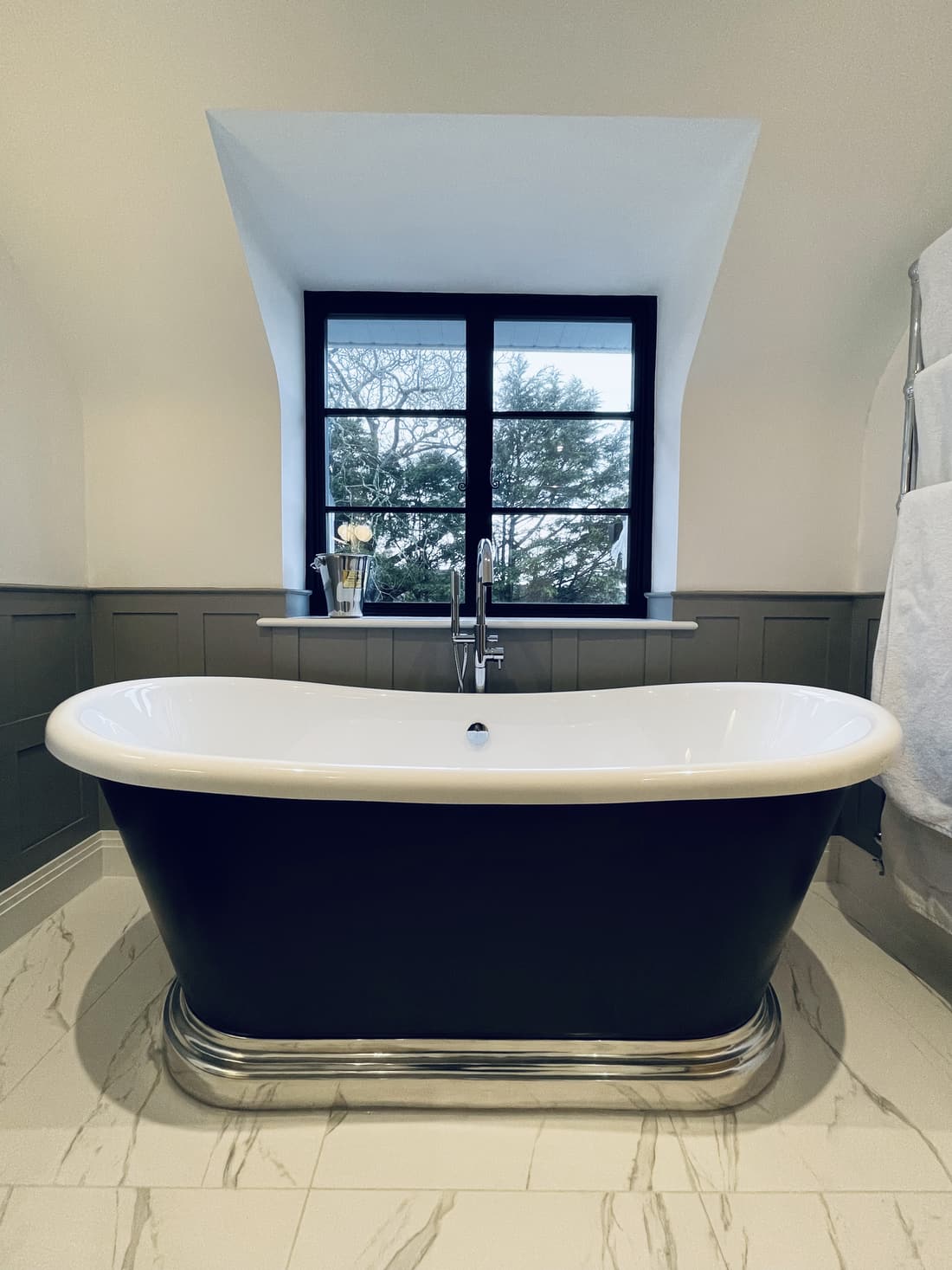 The boat bath head-on in the dormer, white inside and dark outside on a moulded polished metal plinth, a floor-standing chrome mixer beside it, bare branches and evergreen through the black-framed window, and a chrome towel rail at the right.