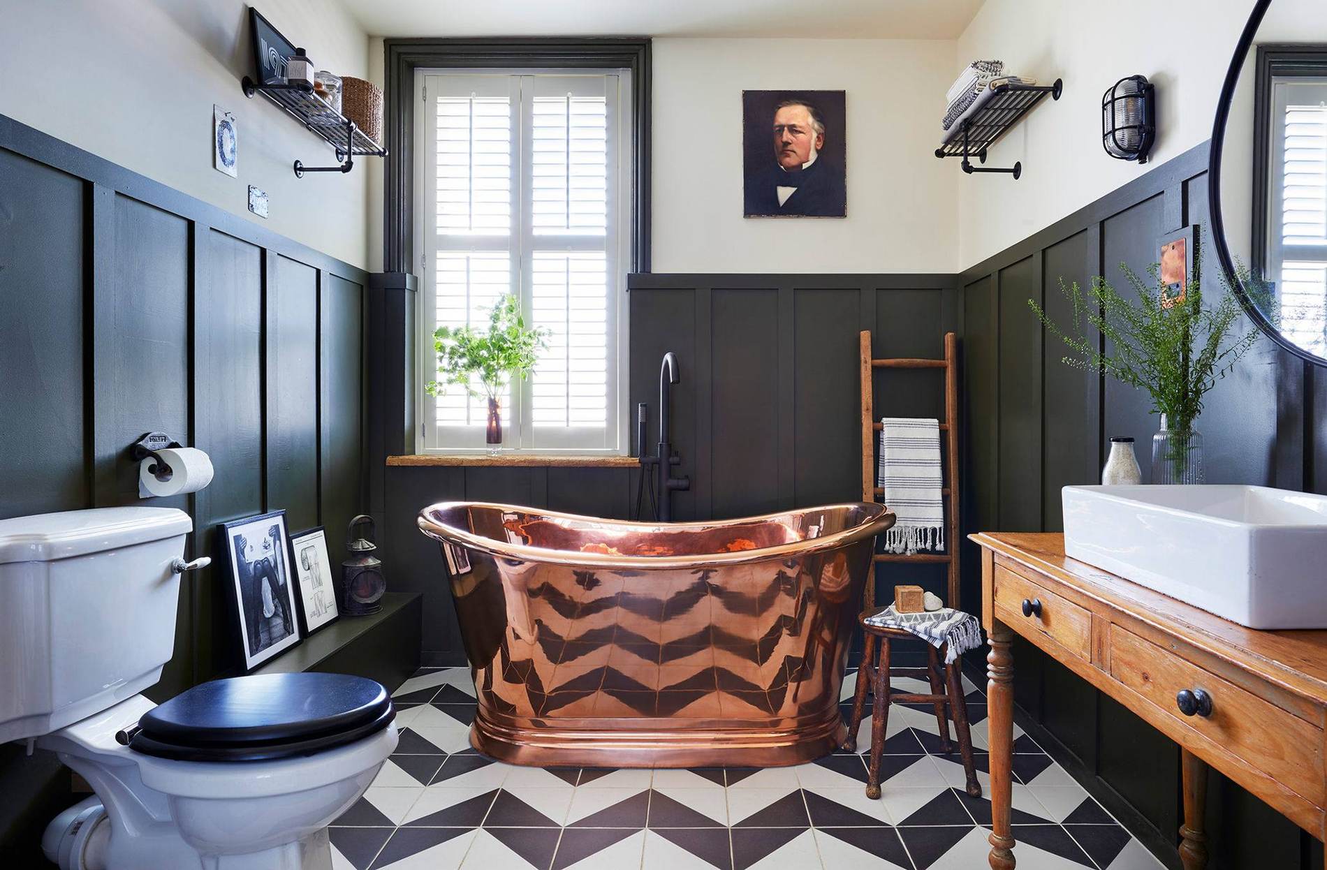 A copper roll-top bath against dark panelled walls, a black and white geometric tiled floor, a shuttered sash window and a framed antique portrait above.