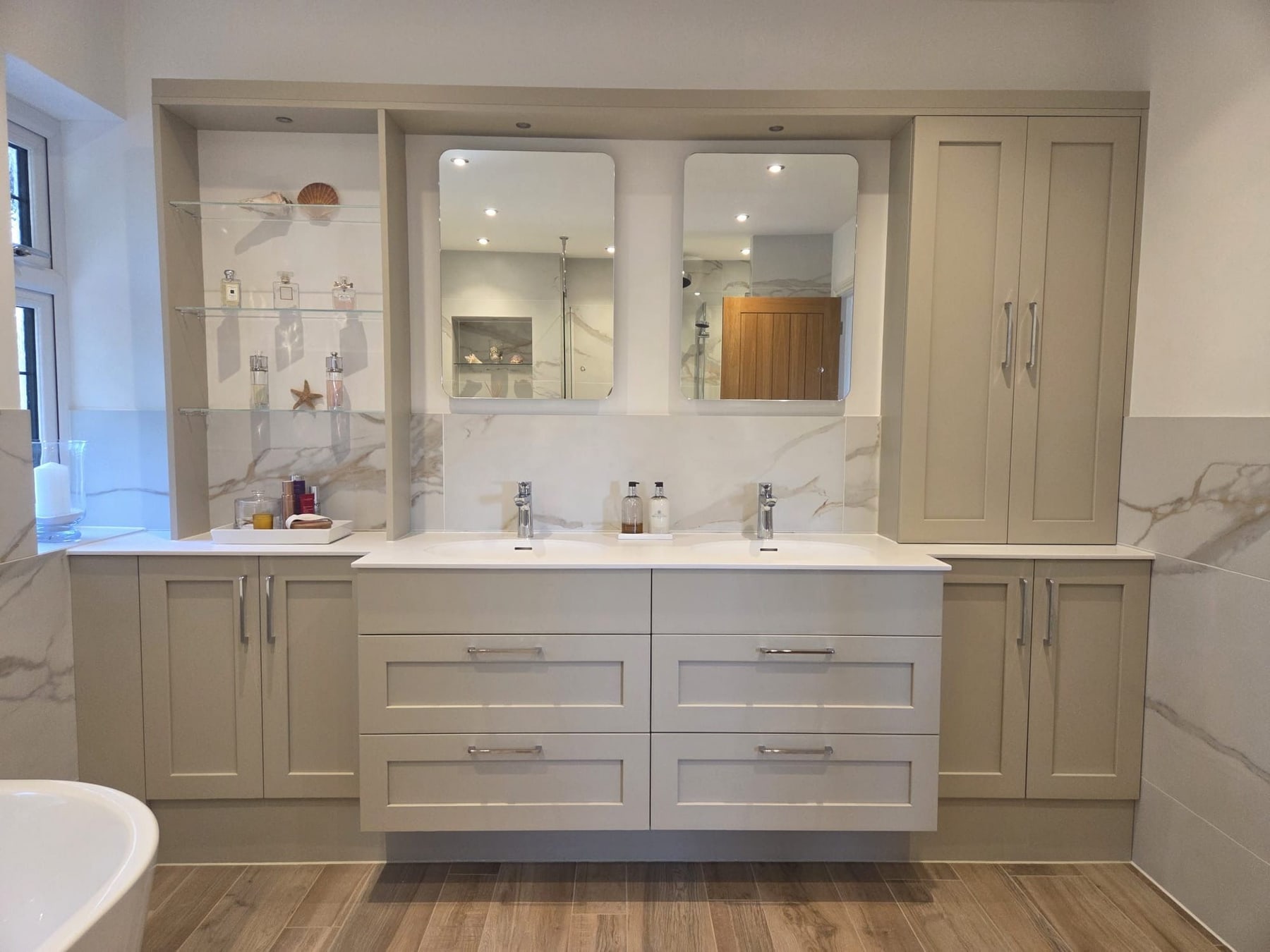 A fitted taupe shaker furniture run with twin basins in a white top, two soft-cornered mirrors, open glass display shelving and a tall cupboard, against marble-effect tiling.