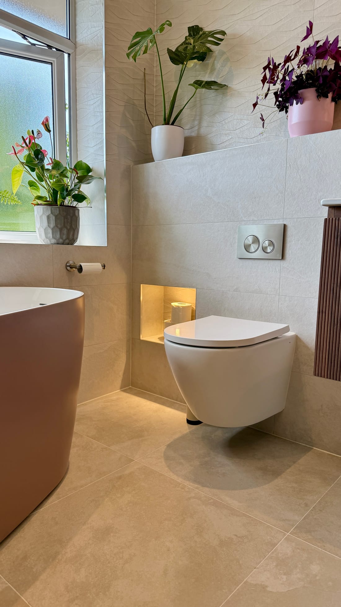The wall-hung WC with a round chrome flush plate above it, the lit recessed niche holding a toilet roll beside it, plants on the tiled ledge and the window sill behind, and the clay-coloured side of the bath at the left.