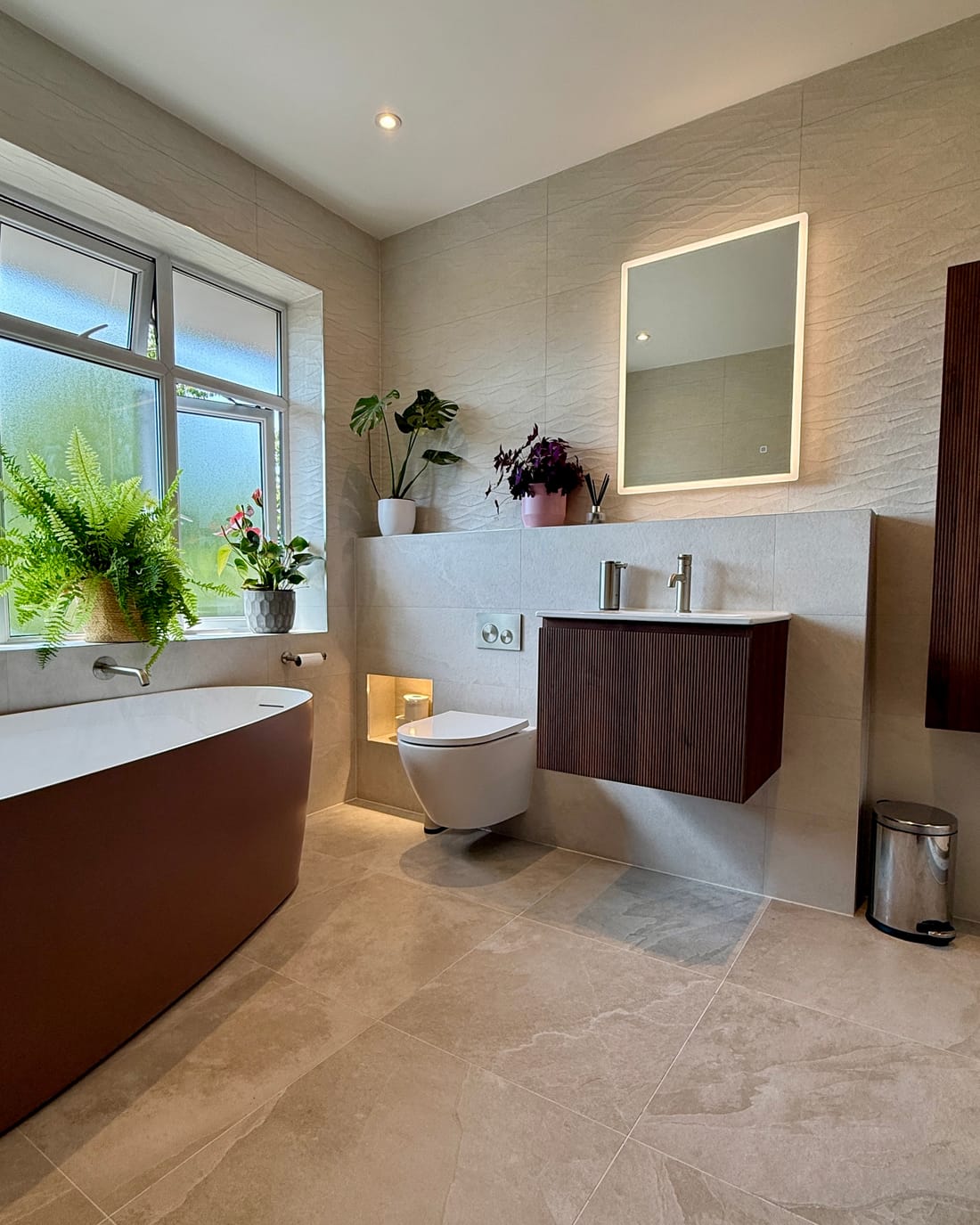 A bathroom in sand-coloured stone-effect porcelain: a freestanding bath with a white interior and a matt clay-coloured outer skin under an obscure-glazed window lined with plants, a wall-hung fluted walnut vanity with a white basin top beneath a backlit rectangular mirror, and a wall-hung WC beside a lit recessed niche.